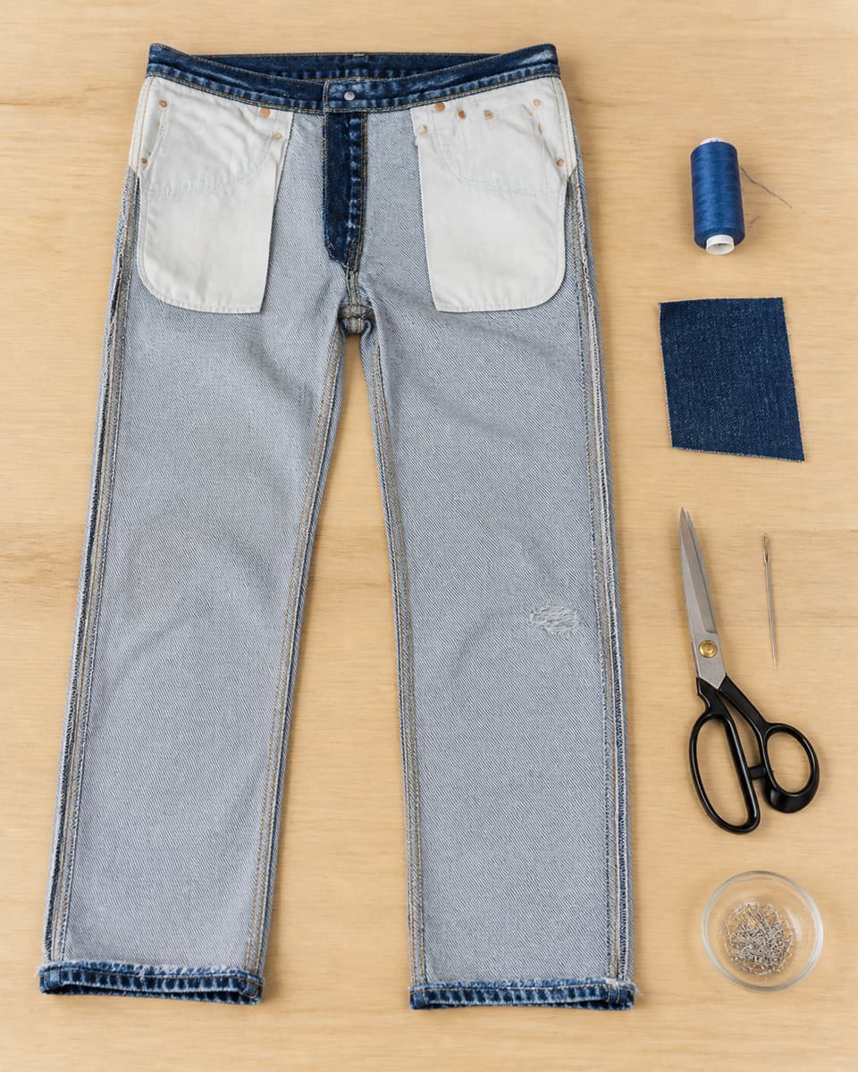 Jean retourné à plat entouré d'une aiguille, d'une bobine de fil, de ciseaux et d'une pièce de denim sur une table claire.