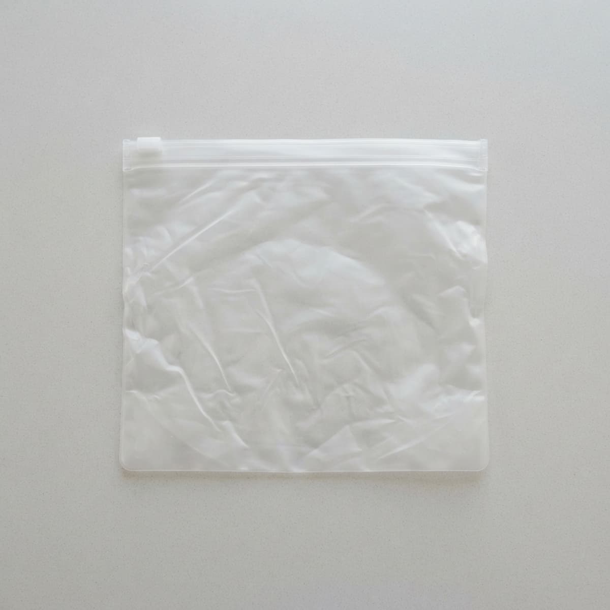 Sac en PEVA fin et translucide, légèrement froissé, avec une fermeture à glissière transparente, posé sur une surface lisse gris clair.